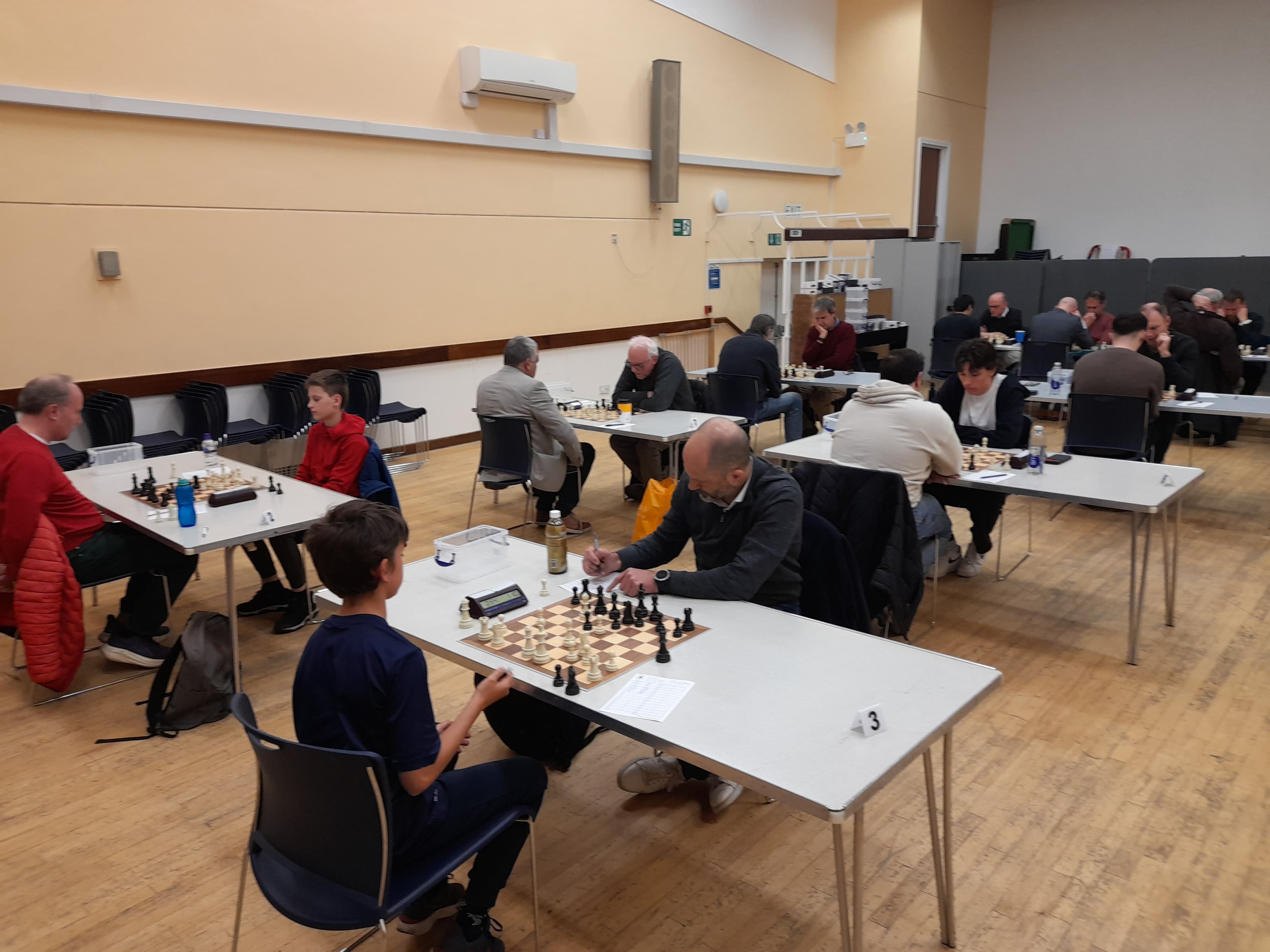 People of various ages compete in a chess tournament at tables in a large hall.